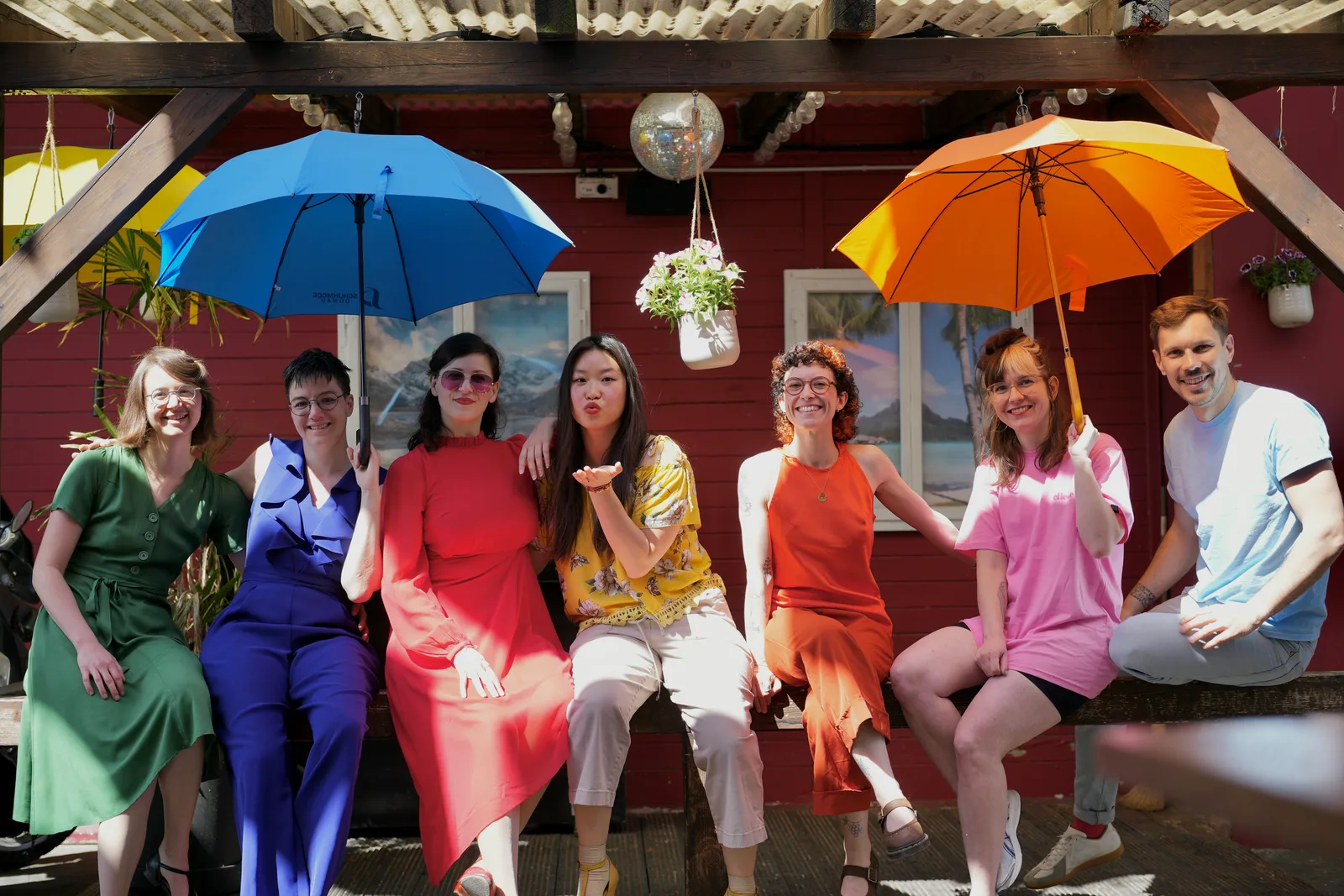 QSXZ team sitting in a row holding rainbow-coloured umbrellas in front of a red house